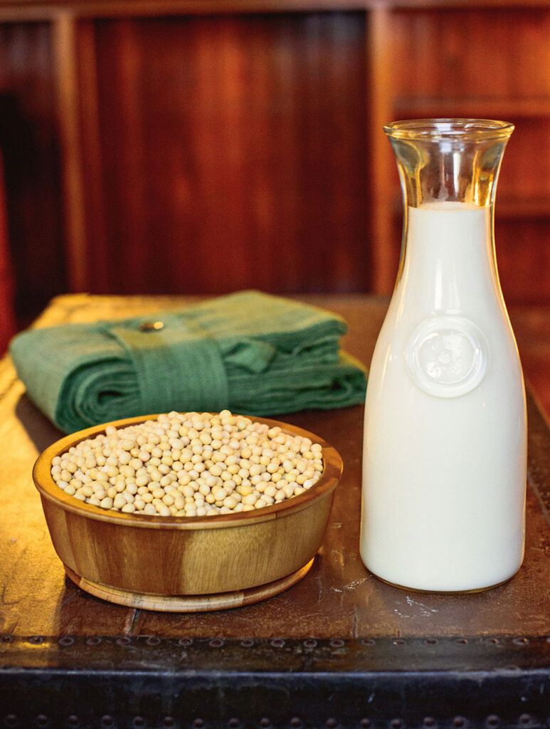 A wooden bowl filled with soybeans a tall glass container of soy milk.