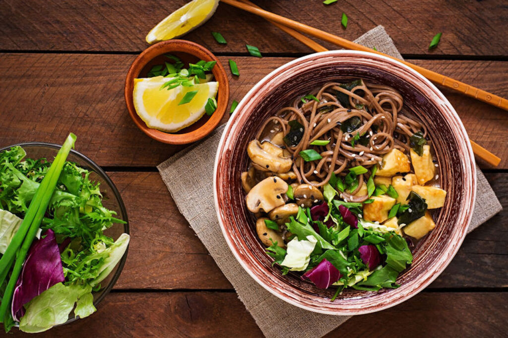 A bowl filled with noodles, mushrooms, tofu, and salad mix on a table next to chopsticks. There's also a bowl with lemon slices and another bowl with more salad mix.