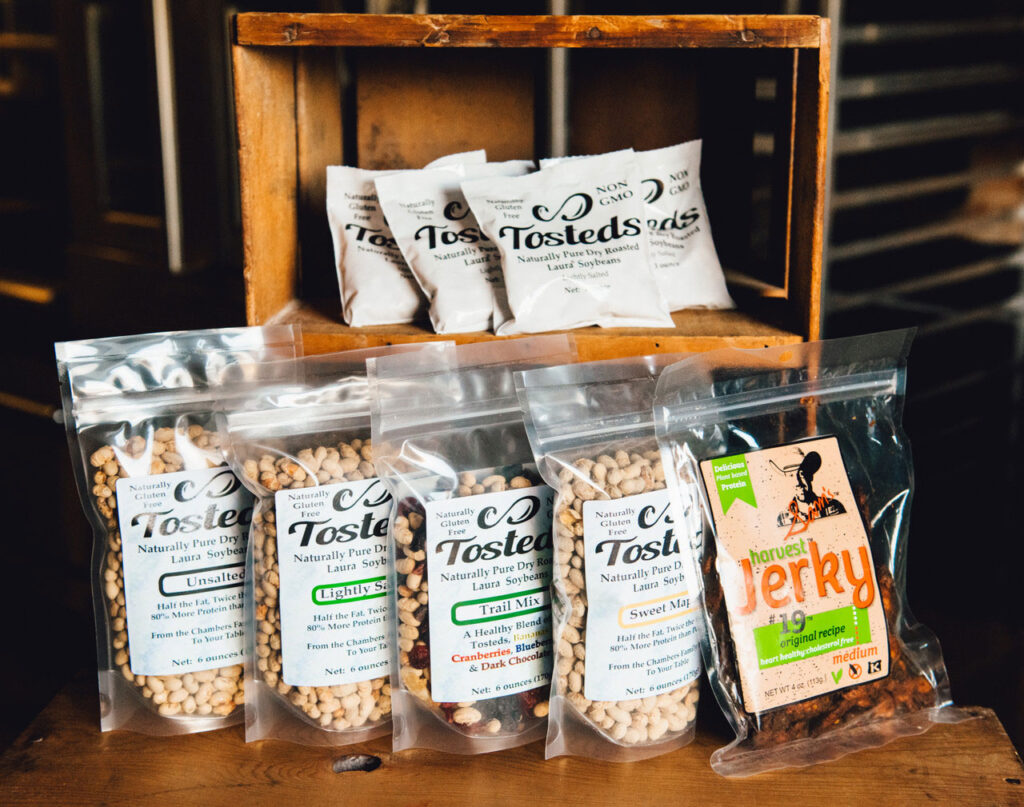 A wooden stand with small bags of Tosteds, with larger resealable clear bags of Tosteds and Tosteds Trail Mix.