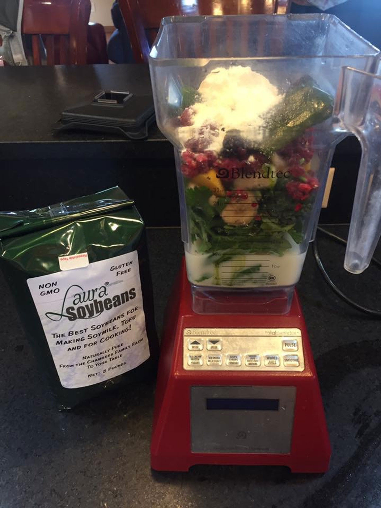 A blender filled with soymilk, coconut milk, greens, some fruit and protein powder. A package of Laura Soybeans sits next to it on the ground.