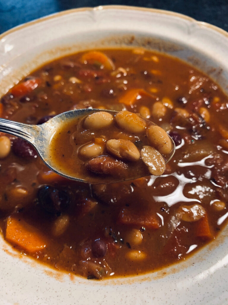 A close up of a spoon over a bowl of vegan chili filled with soybeans and various veggies.