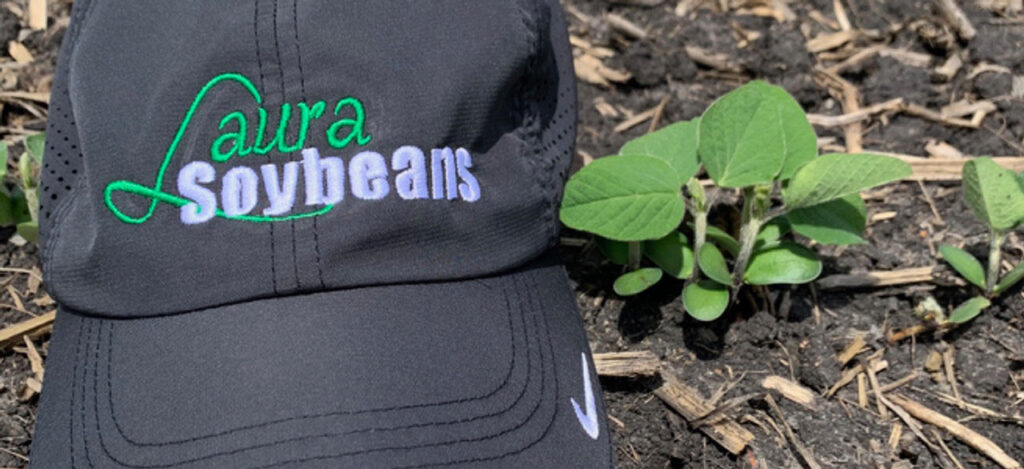 A black and white Laura Soybeans hat next to a small Laura Soybean plant.