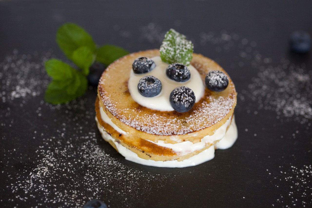 Stacked pancakes with cream between the layers and topped with a dusting of powdered sugar, blueberries, and mint leaf.