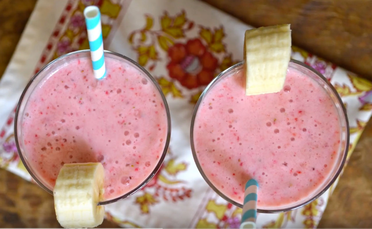 Two glasses with pink-ish smoothies. Each glass has a white and blue striped straw and banana slices set on the rim.