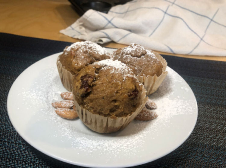 A white plate with 3 cherry almond muffins made with Laura soymilk and a few almonds. The muffins and plate are dusted with powdered sugar.
